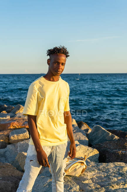 Happy African American male in casual clothes standing on rocky seashore while smiling and looking at camera in summer evening — Stock Photo