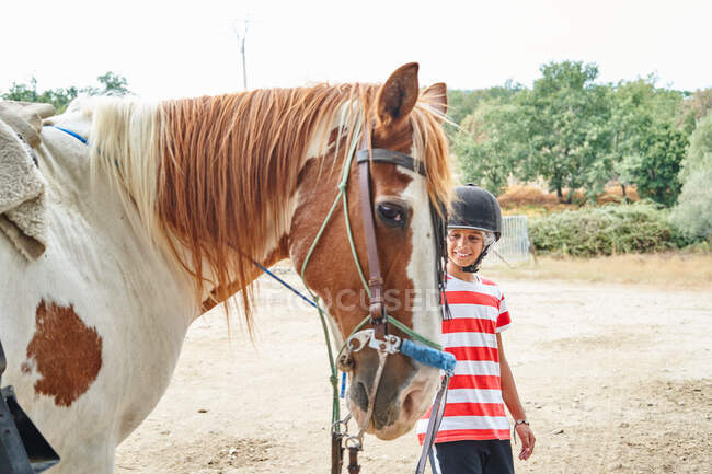 Позитивна дитина в жокейній шапці і повсякденному одязі стоїть біля коня з вуздечкою на піщаному ґрунті біля зелених дерев і рослин у сільській місцевості вдень. — стокове фото