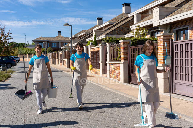 Joven equipo de limpieza en uniforme llevando escobas con cucharas y cubos mientras camina por la calle pavimentada cerca de casas residenciales en un día soleado - foto de stock