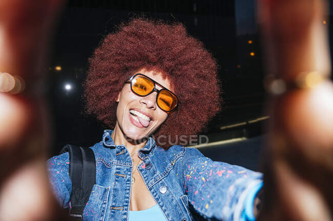 Entzückte Frau mit Afro-Frisur und Sonnenbrille blickt mit herausgestreckter Zunge in die Kamera, während sie abends auf der Straße Selbstporträts macht — Stockfoto