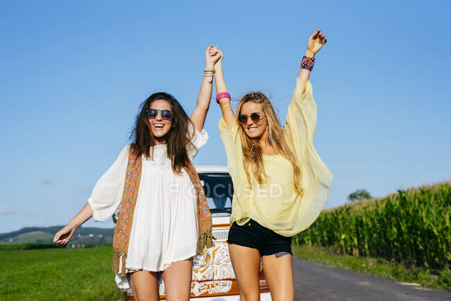 Dos hermosas y felices chicas caucásicas vestidas con ropa de verano de pie en la carretera fuera de una furgoneta - foto de stock