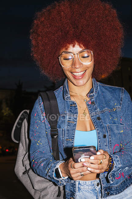 Веселая женщина с прической афро в модном джинсовом костюме, отправляющая сообщения по мобильному телефону, стоя на улице в вечернее время — стоковое фото