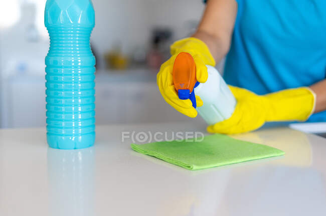 Анонимная женщина в жёлтых резиновых перчатках распыляет моющее средство на ткань при уборке светлой кухни — стоковое фото