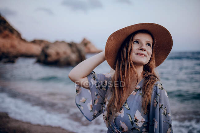Счастливая женщина в стильном платье и шляпе стоя, глядя в сторону и подняв руку на берегу моря в летний вечер — стоковое фото