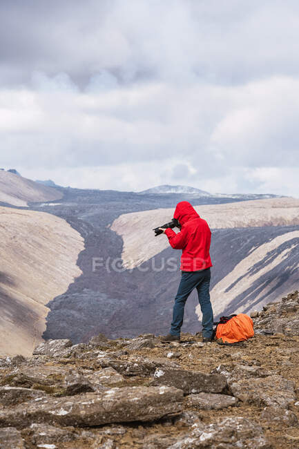 Vista lateral do fotógrafo masculino em roupa exterior em pé no topo do penhasco rochoso perto do vulcão ativo Fagradalsfjall com lava preta na Islândia durante o dia — Fotografia de Stock