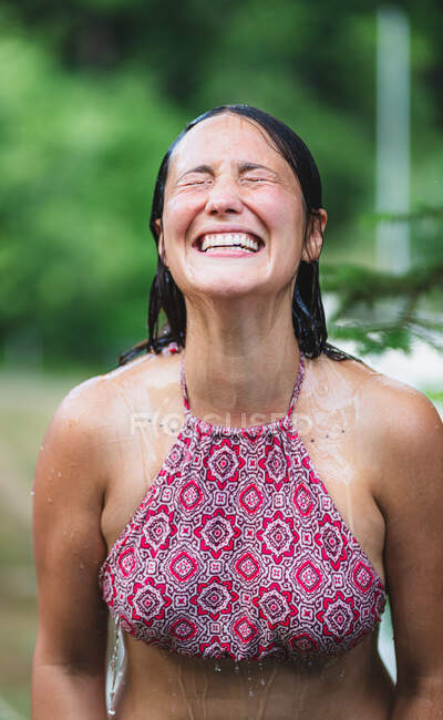 Happy female standing with closed eyes in top and pouring water from bottle on head against green trees in park in summer time — Stock Photo