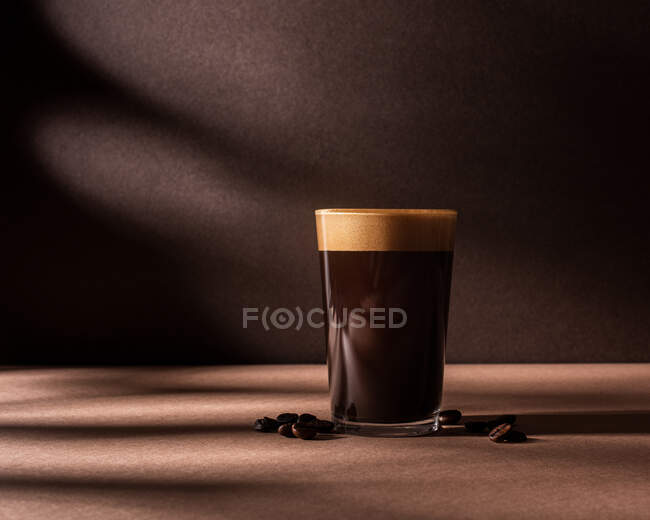Vista frontal de una taza de café negro con una capa de espuma y colocado junto a algunos granos de café en una mesa marrón y la pared con sombras oscuras - foto de stock