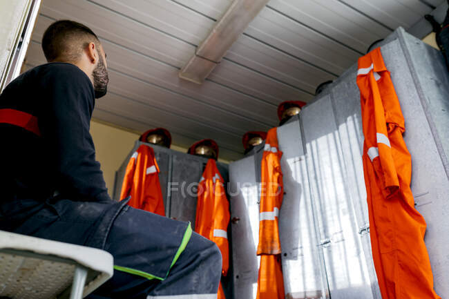 Desde abajo bombero desconocido sentado cerca de una fila de armarios de metal con cascos en la parte superior y uniforme naranja colgando - foto de stock