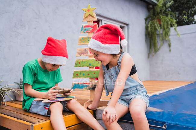 Дети в красных шляпах Санты просматривают мобильный телефон, сидя рядом с декоративной окрашенной елкой в светлом помещении во время празднования праздника — стоковое фото