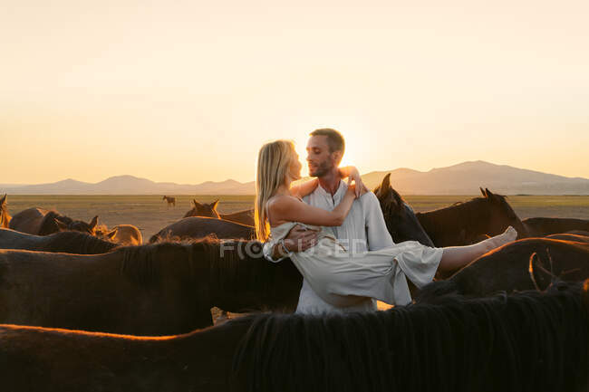 Homem segurando bela namorada de cabelos entre cavalos no campo pasto enquanto olham um para o outro — Fotografia de Stock