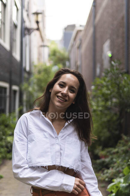 Lächelnd Attraktive Frau in Freizeitkleidung, die in der Nähe von Gebäuden und Pflanzen auf einer schmalen Straße spaziert und dabei in die Kamera blickt — Stockfoto