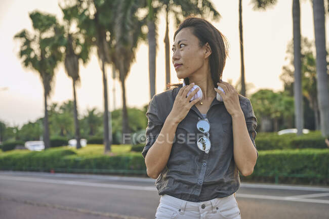 Серьёзная азиатка в современных белых наушниках смотрит вдаль, стоя рядом с дорогой на улице города с зелеными деревьями — стоковое фото