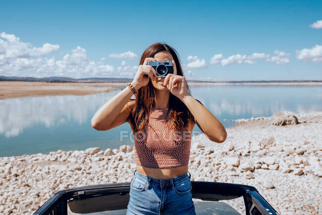 Young female standing on car while taking picture on old fashioned photo camera on coast of blue pond — Stock Photo
