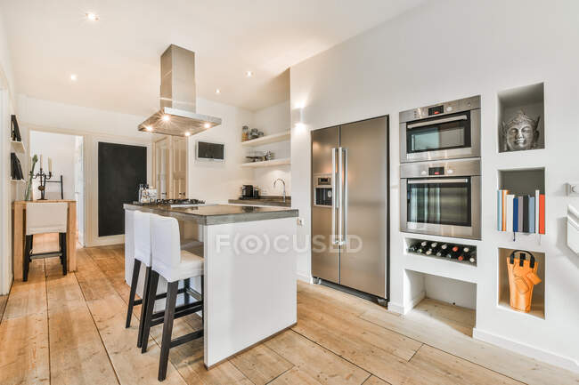 Современная кухня со столом и вытяжкой против холодильника и электрической печи в светлом доме с деревянным полом — стоковое фото