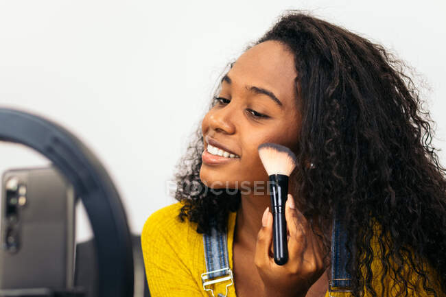 Femme afro-américaine positive utilisant un pinceau pour appliquer des cosmétiques sur la joue tout en enregistrant une vidéo pour vlog beauté — Photo de stock
