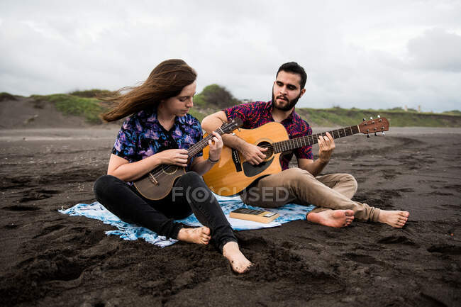 Homme souriant jouant de la guitare acoustique avec une amie positive jouant de l'ukulélé assis sur une côte sablonneuse dans la nature par temps nuageux — Photo de stock
