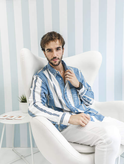Elegant bearded guy looking at camera and adjusting collar of t shirt while sitting on white armchair — Stock Photo