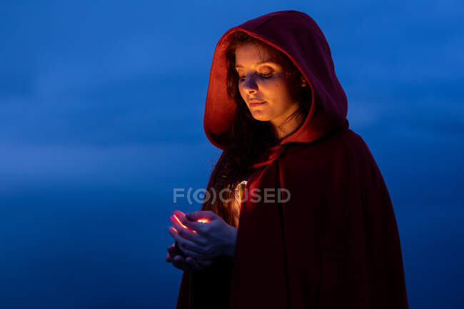 Serious female in Victorian styled outfit with red cape looking down while standing with glowing candle in hands in dark nature — Stock Photo