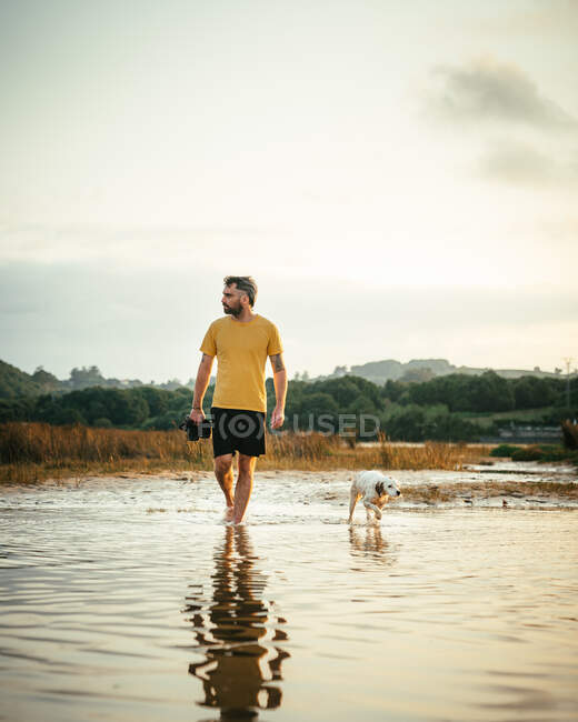 Full body of male owner with boots in hands walking in water near running dog on summer day in nature — Stock Photo