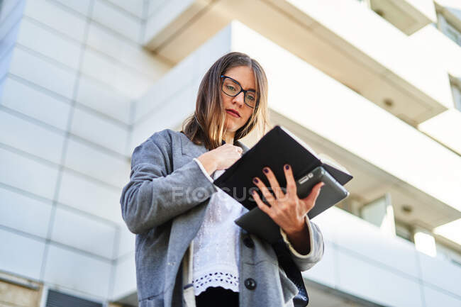 Снизу задумчивая молодая предпринимательница в очках, стоя с планшетом и ноутбуком и думая о планах — стоковое фото