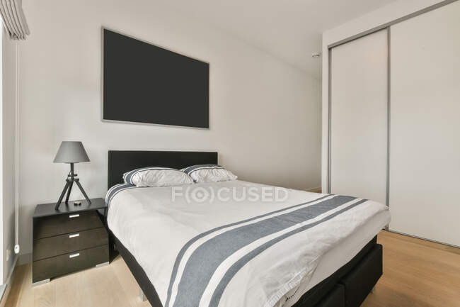 Удобная кровать с постельным бельем на стене с тумбочкой и телевизором в стильной светлой спальне со шкафом в квартире — стоковое фото