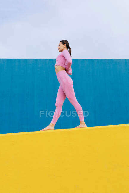 Vista laterale di tutta la lunghezza della donna a piedi nudi sottile praticare yoga in preghiera inversa con le mani dietro la schiena sulla parete gialla e blu — Foto stock