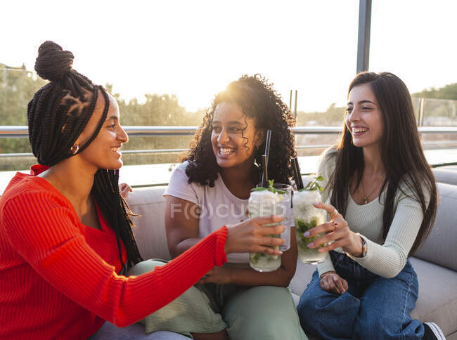 Joyeuses amies multiraciales dans des vêtements à la mode assises sur un canapé confortable et des verres de mojito alcoolisé tout en passant du temps ensemble sur la terrasse au soleil — Photo de stock