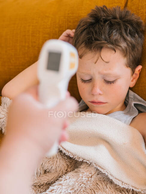 De arriba de la cosecha el padre con el termómetro infrarrojo que mide la temperatura del chiquitín triste enfermo tendido bajo la manta en el sofá en casa - foto de stock