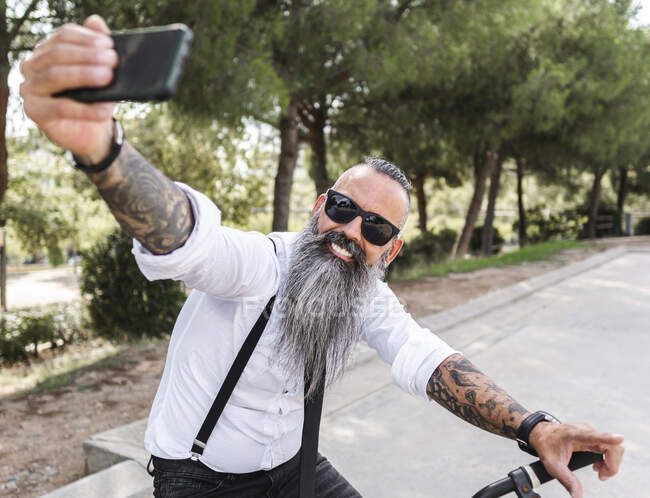 Positiv bärtiger Mann mit Sonnenbrille und weißem Hemd, der sich auf dem Smartphone porträtiert, während er im Park mit Bäumen auf dem Fahrrad sitzt — Stockfoto