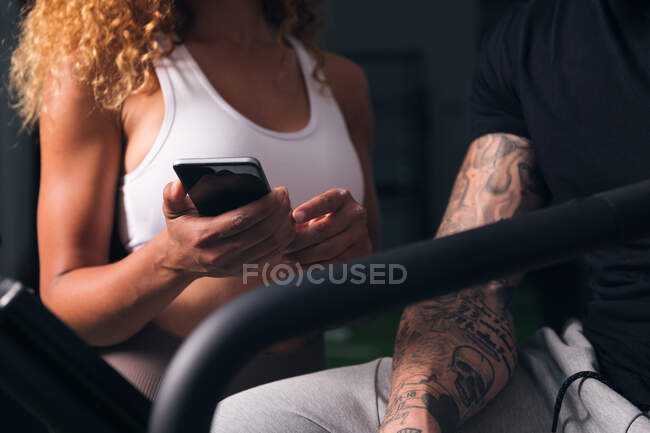 Cortar hembra irreconocible con el pelo rizado en la parte superior blanca demostrando smartphone a macho con tatuajes en la habitación - foto de stock
