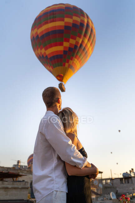 Low angle of anonymous embracing couple standing on street and looking at hot air balloons floating in evening cloudless sky — Stock Photo