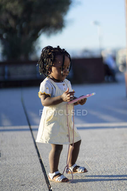 Vista lateral da calma menina afro-americana com tranças pretas em roupas elegantes de pé com smartphone em caso colorido em mãos na passarela de asfalto na rua no dia ensolarado — Fotografia de Stock