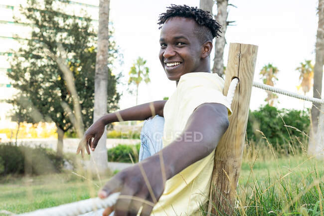 Счастливый афроамериканец в повседневной одежде с короткими волосами, сидящий на траве с рукой на согнутой ноге и опирающийся на веревочный забор — стоковое фото