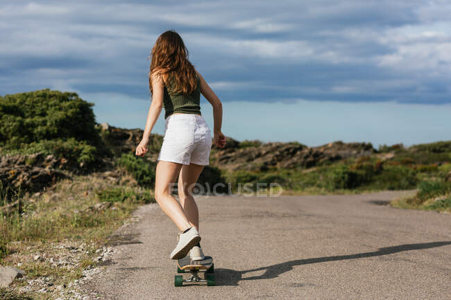 Corpo inteiro visão traseira de irreconhecível jovem fêmea montando longboard na estrada de asfalto entre colinas sob céu nublado — Fotografia de Stock