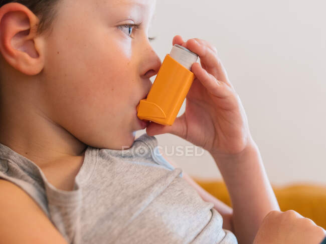 Vista lateral del niño enfermo contenido usando respirador y oxígeno respiratorio del inhalador mientras está sentado en el sofá en casa - foto de stock