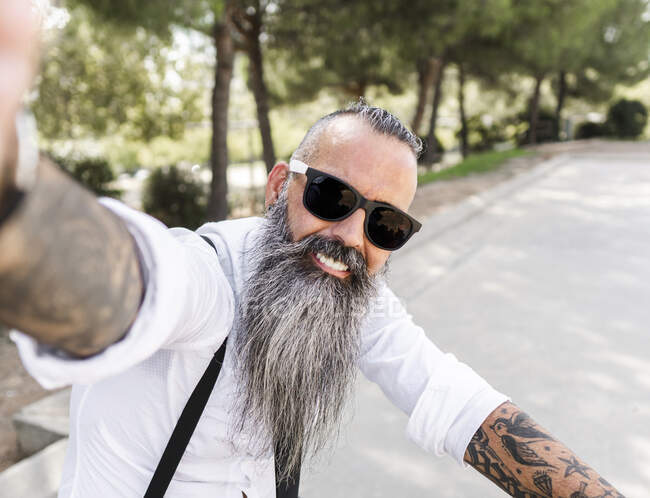 Позитивний бородатий чоловік в сонцезахисних окулярах і білій сорочці, взявши власний портрет на смартфон, сидячи на велосипеді в парку з деревами — стокове фото
