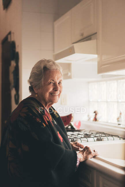 Visão lateral de feliz positivo sênior fêmea com cabelos grisalhos vestindo roupas quentes em pé na pia na cozinha — Fotografia de Stock