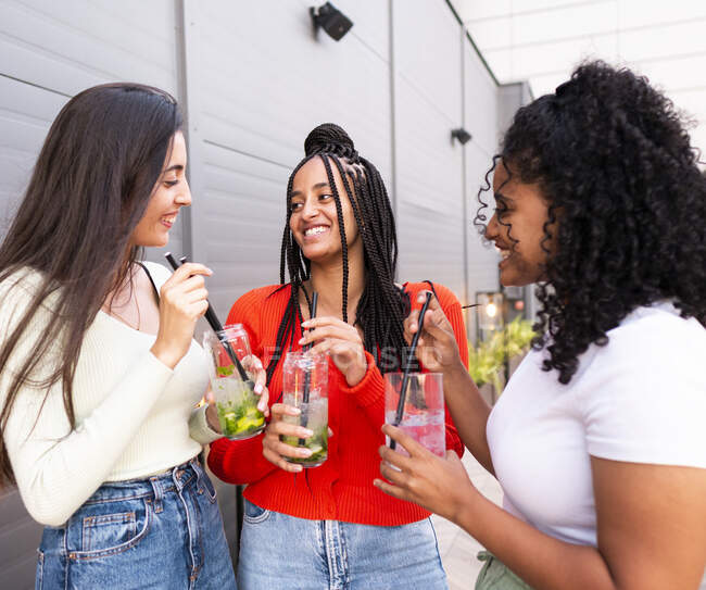 Cheerful multiracial women in trendy clothes standing and drinking glasses of mojito against gray wall in daytime — Stock Photo