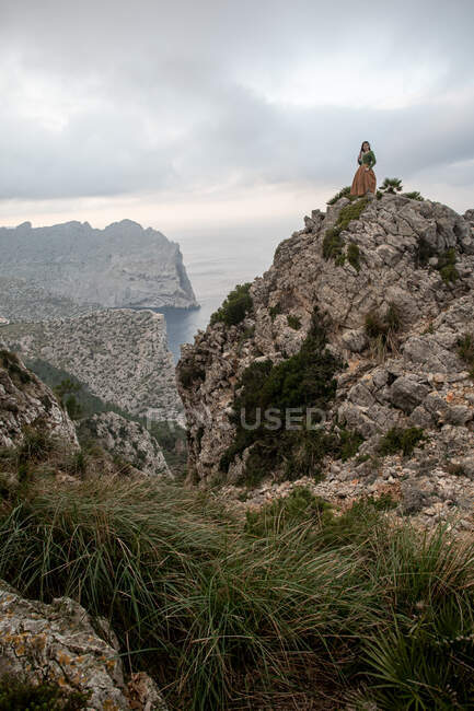 Full body side view of dreamy female in old fashioned clothes standing on edge of stony cliff near sea in misty weather — Stock Photo