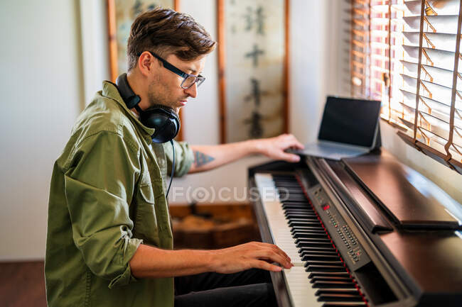 Вид сбоку мужчины-музыканта в повседневной одежде, играющего на фортепиано, сидя в современной квартире у окна — стоковое фото