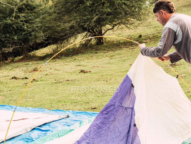 Seitenansicht eines jungen männlichen Reisenden in Freizeitkleidung, der sein Zelt auf einer grünen, grasbewachsenen Wiese öffnet, während er in der Natur unterwegs ist — Stockfoto