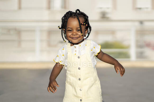 Веселая афроамериканская девочка с косичками в стильной одежде, стоящая на улице напротив здания в солнечный день — стоковое фото