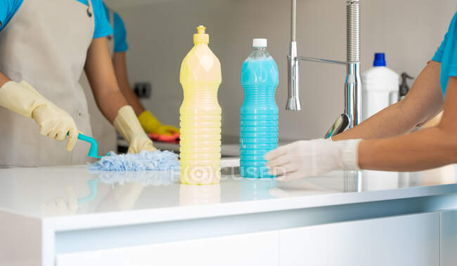 Crop anonymous cleaner in yellow rubber gloves with detergent bottles cleaning light kitchen — Stock Photo