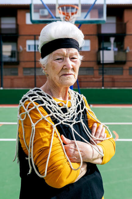 Женщина в спортивной одежде и белой сетке бокового вида смотрит в камеру, стоя на спортивной площадке с баскетбольным обручом во время тренировки — стоковое фото