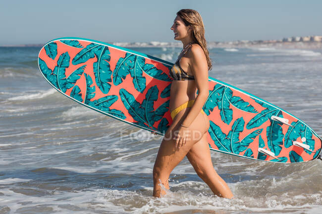 Vista lateral de una surfista deportiva con tabla de surf paseando en el mar ondulado durante el entrenamiento en un resort tropical en un día soleado - foto de stock