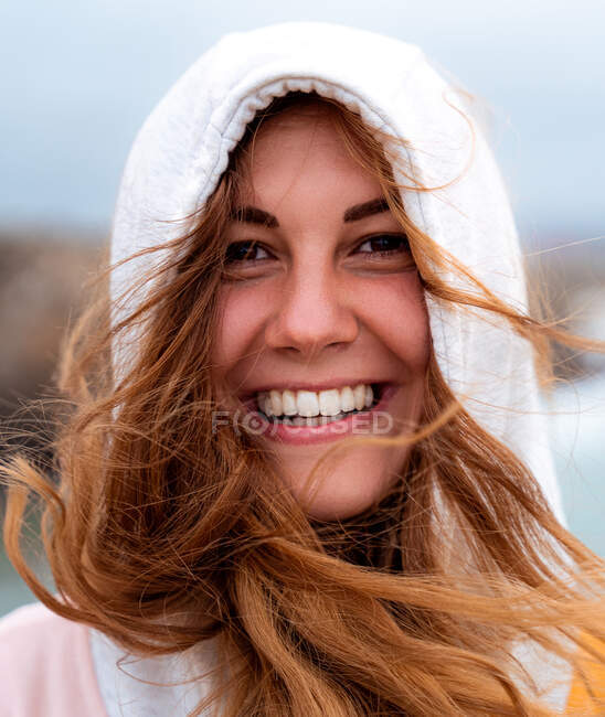 Portrait de charmante femme énigmatique gaie avec des cheveux gingembre volant et à capuche regardant la caméra par jour venteux en Galice — Photo de stock
