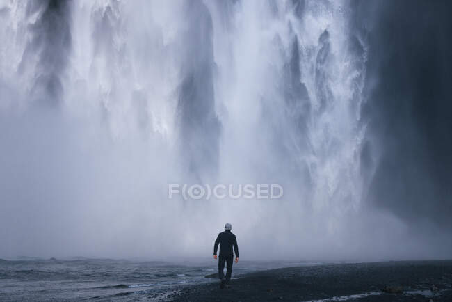 Обратный вид на анонимного исследователя мужского пола в повседневной одежде и шляпе, идущего возле дикой реки с черным песчаным берегом к мощному водопаду Скогафосс во время путешествия по Исландии — стоковое фото