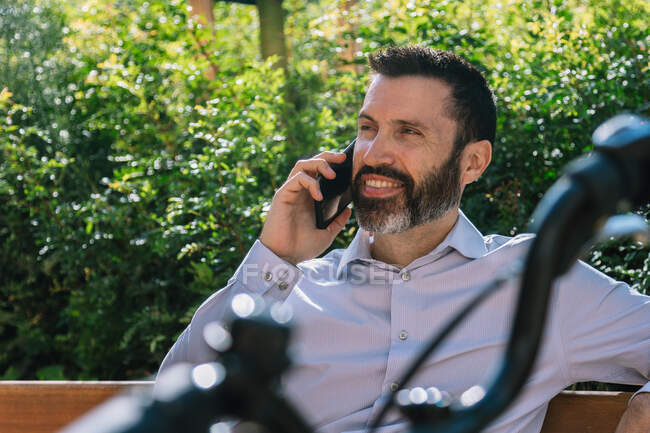 Чоловічий працівник в офіційному одязі сидить у лавці в парку з велосипедом і розмовляє на мобільному телефоні — стокове фото