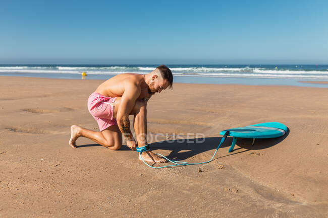 Visão lateral do corpo inteiro do macho ativo em shorts de natação anexando corda da perna enquanto se prepara para surfar na praia de areia perto do mar — Fotografia de Stock