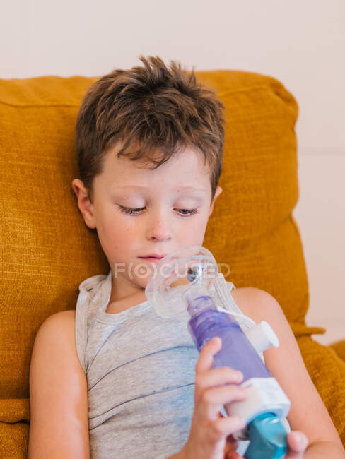Conteúdo menino doente usando respirador e respirando oxigênio do inalador enquanto sentado no sofá em casa — Fotografia de Stock